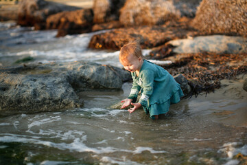 Toddler girl in a dress plays with water on the seashore at sunset, sand and stones, lifestyle