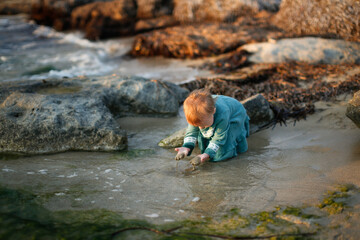 Toddler girl in a dress plays with water on the seashore at sunset, sand and stones, lifestyle