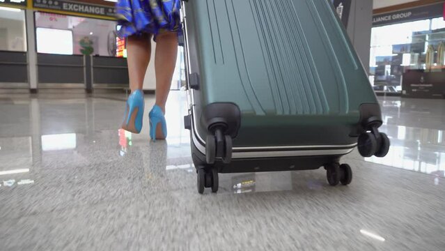 Close-up of female traveler's feet in high-heeled shoes at an empty airport movement of women's feet in blue shoes and blue dress carrying traveling luggage on wheels