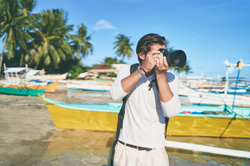 Photography and travel. Young man with rucksack taking photo with his camera on the sea beach near fishing boats.