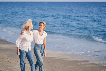 Outdoor portrait of smiling happy caucasian senior mother with her adult daughter hugging and walking on sea beach. - Powered by Adobe