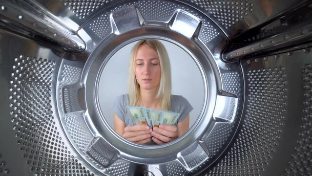 Young beautiful housewife counts dollars of money near washing machine. View from inside on wide gogol. Woman washes or saves on washing clothes. Saving family budget. Debts and creldites. Service