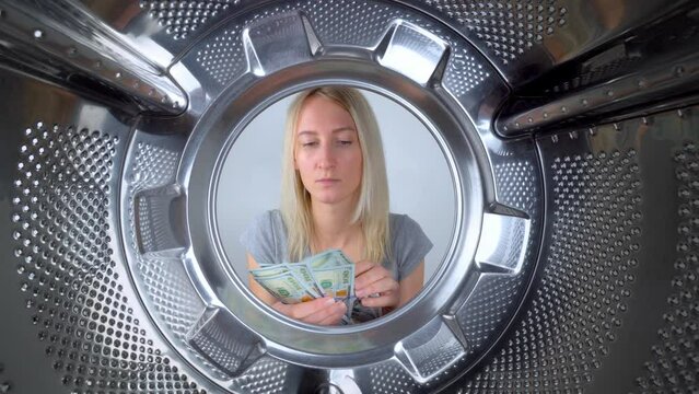 Young beautiful housewife counts dollars of money near washing machine. View from inside on wide gogol. Woman washes or saves on washing clothes. Saving family budget. Debts and creldites. Service