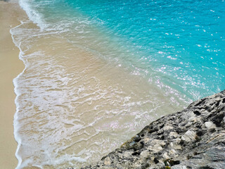 Sunny seascape with turquoise wave hitting white beige sandy coast and grey limestone rock. Colorful sea waters with white foam on sandy beach. Rock, sand and sea