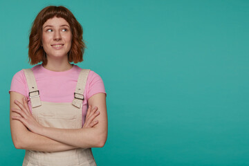 indoor portrait of young ginger female posing over blue background looking aside at copy space with smile on her face