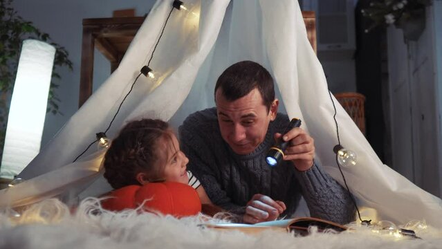 happy family at home dream. dad and daughter happy family at home reading a book in lockdown the evening in stay home homemade wigwam house. kid dream coronavirus concept. dad daughter read book
