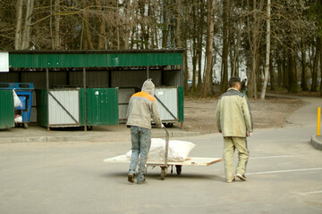 Two workers in work clothes carry garbage to the garbage on a cart. Cloudy day, outskirts of the city. Rear view.