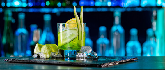 Cocktail with cucumber, ice on bar counter in a restaurant, pub. Fresh tonic drink with lime juice, mint, gin, cucumber juice. Alcoholic cooler beverage at nightclub on dark background - obrazy, fototapety, plakaty