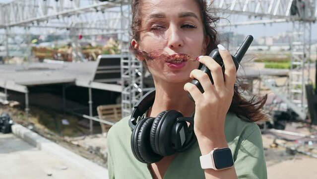 Event manager portrait. Installation of stage equipment and preparing for a live concert open air. Summer music city festival. Young serious woman speaks the walkie-talkie.