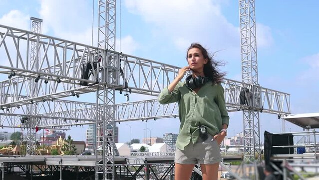 Installation of stage equipment and preparing for a live concert open air. Event manager portrait. Summer music city festival. Young serious woman speaks the walkie-talkie.