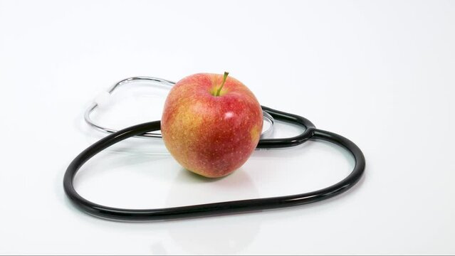 A stethoscope wraps a ripe red apple on a white background. The concept of health care, healthy food, diet, disease prevention. Stop motion animation.