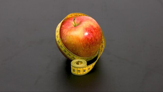 A red ripe apple wrapped in a yellow tailor's measuring tape on a black background. The concept of healthy food, diet, weight loss, . Stop motion animation.