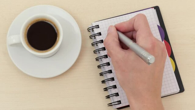 Man writes a travel checklist in notepad on table next to white coffee cup with espresso