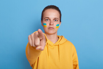 Indoor shot of serious patriotic woman wearing hoodie with blue and yellow flag on cheeks, looking and pointing finger to camera, Russian you can stop the war, posing isolated over blue background.
