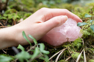 Hand holding a rose quartz crystal or raw gemstone minerals on a forest floor with lush greenery. 