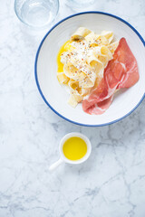 Creamy pappardelle pasta with prosciutto and poached egg in a white plate, above view on a light-grey marble background, vertical shot with space