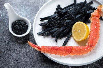 Closeup of black pasta with black caviar and boiled kamchatka crab leg served on a white plate,...