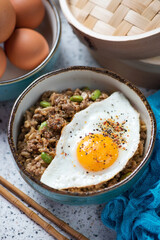 Bowl of fried rice and minced meat topped with a chicken egg, vertical shot on a beige granite background, middle closeup