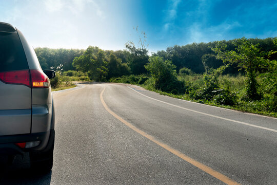 Rear side of family car drving on the asphalt road with turn on signal. Road path that leads forward and has a curving path. Backgroun of green grass and trees under the blue sky.