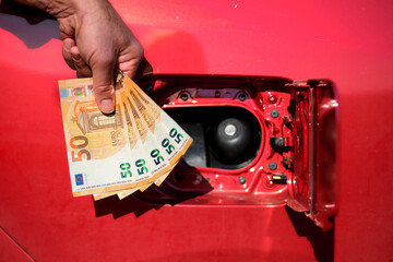 A hand holding many fifty euro notes next to a car's fuel tank, symbolizing the raising fuel and...