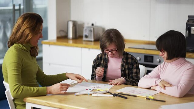 Two girls with down syndrome draw at the kitchen next to the teacher or mother