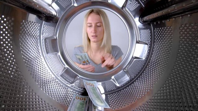 Young beautiful housewife counts dollars of money near washing machine. View from inside on wide gogol. Woman washes or saves on washing clothes. Saving family budget. Debts and creldites. Service