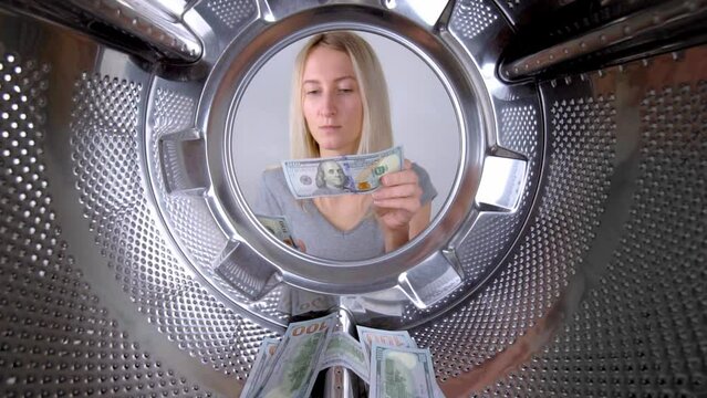 Young beautiful housewife counts dollars of money near washing machine. View from inside on wide gogol. Woman washes or saves on washing clothes. Saving family budget. Debts and creldites. Service