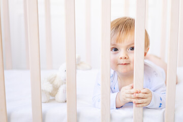 laughing baby blonde boy lies in a crib behind the side in a crib at home with blue and white cotton bedding and smiles in the morning, the concept of children's goods and accessories