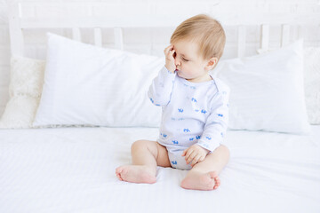 a blonde baby boy rubs sore inflamed eyes or wants to sleep on a white crib while sitting in a room at home with white cotton bedding, the concept of children's goods and accessories