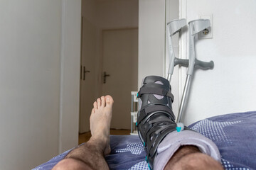 European man after Achilles tendon rupture operation is back home with special physiotherapy shoe and crutches for recovery at home with healthy medicine painkiller drug pills against the hurting leg