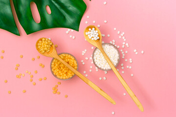 Depilatory wax granules in glass cups and wooden spoons on a pink background.
