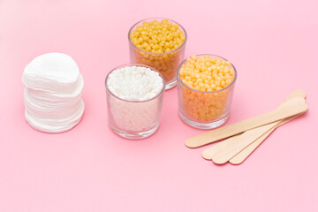 Depilatory wax granules in glass cups and wooden spatulas on a pink background.
