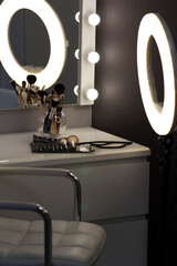 Mirror with light bulbs and ring lamp, near cosmetic brushes on dressing table indoors in makeup room. Shooting in a low key