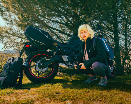 Chica motera viajando por la naturaleza, chica con una c&aacute;mara de fotos junto a una moto negra, viajera fot&oacute;grafa n&oacute;mada 