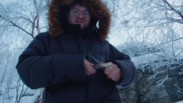 A man with glasses and a dark jacket makes a wooden spear with a knife in the winter forest for his survival.