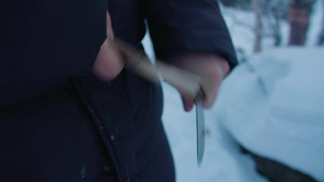A man in the dark jacket makes a wooden spear with a knife in the winter forest for his survival. Close-up.