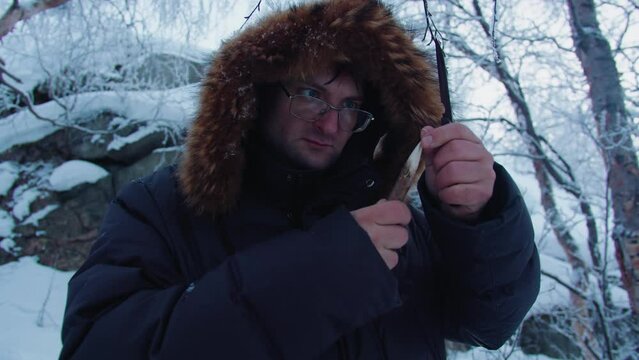 A man with glasses and a dark jacket made a wooden spear with a knife in the winter forest for his survival. He carefully examines the resulting spear.