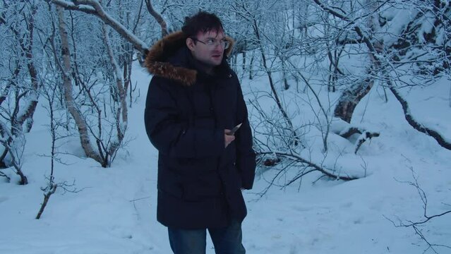 A man in glasses and a dark jacket walks through the winter forest with a wooden spear in his hand and looks warily from side to side.