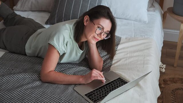 Young happy woman using laptop, typing, smiling and looking at screen, lying on bed at home