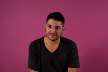 Obraz premium looking at the camera with a shy smile , young brunet man, 20s, wears a blue shirt looking at camera smiling isolated on light pink background studio portrait. People emotions lifestyle concept
