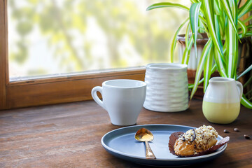 white espresso coffee cup, fresh baked eclair sweet dessert on plate kitchen table against window,...