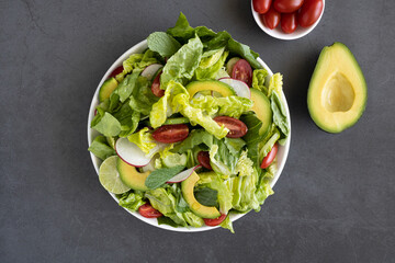 Vegetable diet salad with juicy cherry tomatoes, cucumber, radish and omega-3 rich avocado on a black background, top view. A bowl of crispy green salad with vegetables.