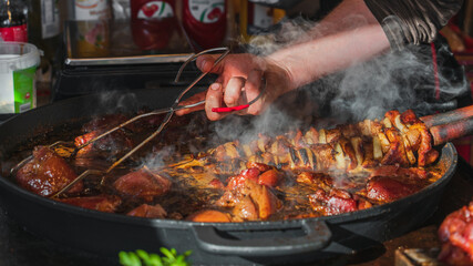 A chef uses metal kitchen tongs to stir pork knuckles in a large frying pan in an open-air street restaurant. Hot white steam over a traditional Polish dish