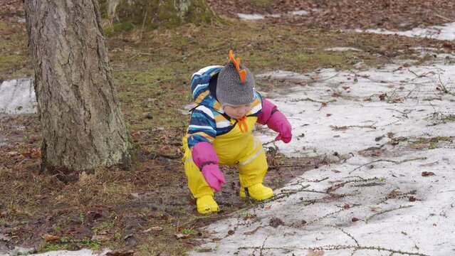 Cute child in yellow waterproof rain pants bibs for kids and rubber boots on a walk in park in early spring, happy kid having fun playing in puddle in cold season. High quality 4k footage