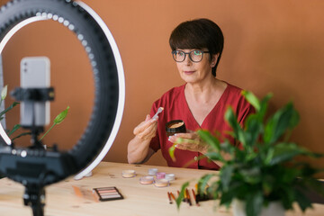 Middle-aged woman talking on cosmetics with makeup eye shadows and blush palette and brushes while recording her video. Mature female making video for her blog on cosmetics