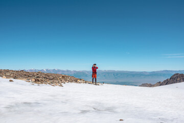 Tourist in red walks on snow mountain near abyss edge on high altitude under blue sky in sunny day. Man with camera on snowy mountain near precipice edge with view to large mountain range in away.