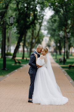 A handsome groom in a formal suit and a lovely bride in a long white dress with a beautiful dress and a bouquet in her hands. The groom hugs the howling bride outdoors.Beautiful wedding photo session.