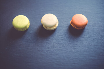 A dark, moody image of chocolate, caramel, and vanilla macaroons arranged on a slate tile, atop a dark grey concrete textured surface background.