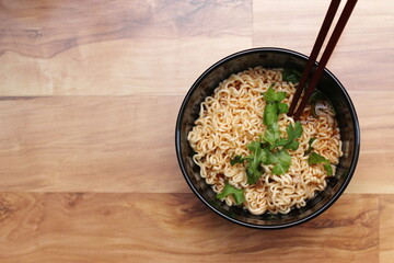 Instant noodles in a black bowl with coriander sprinkles served with chopsticks on a light wooden table.Top view.