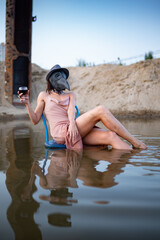 Sexy brunette with short hair, long slender legs and a sporty figure in a silk soft pink wet dress, hat and mask of a plague doctor is sitting on a chair in a lake with a glass of wine - surrealism
